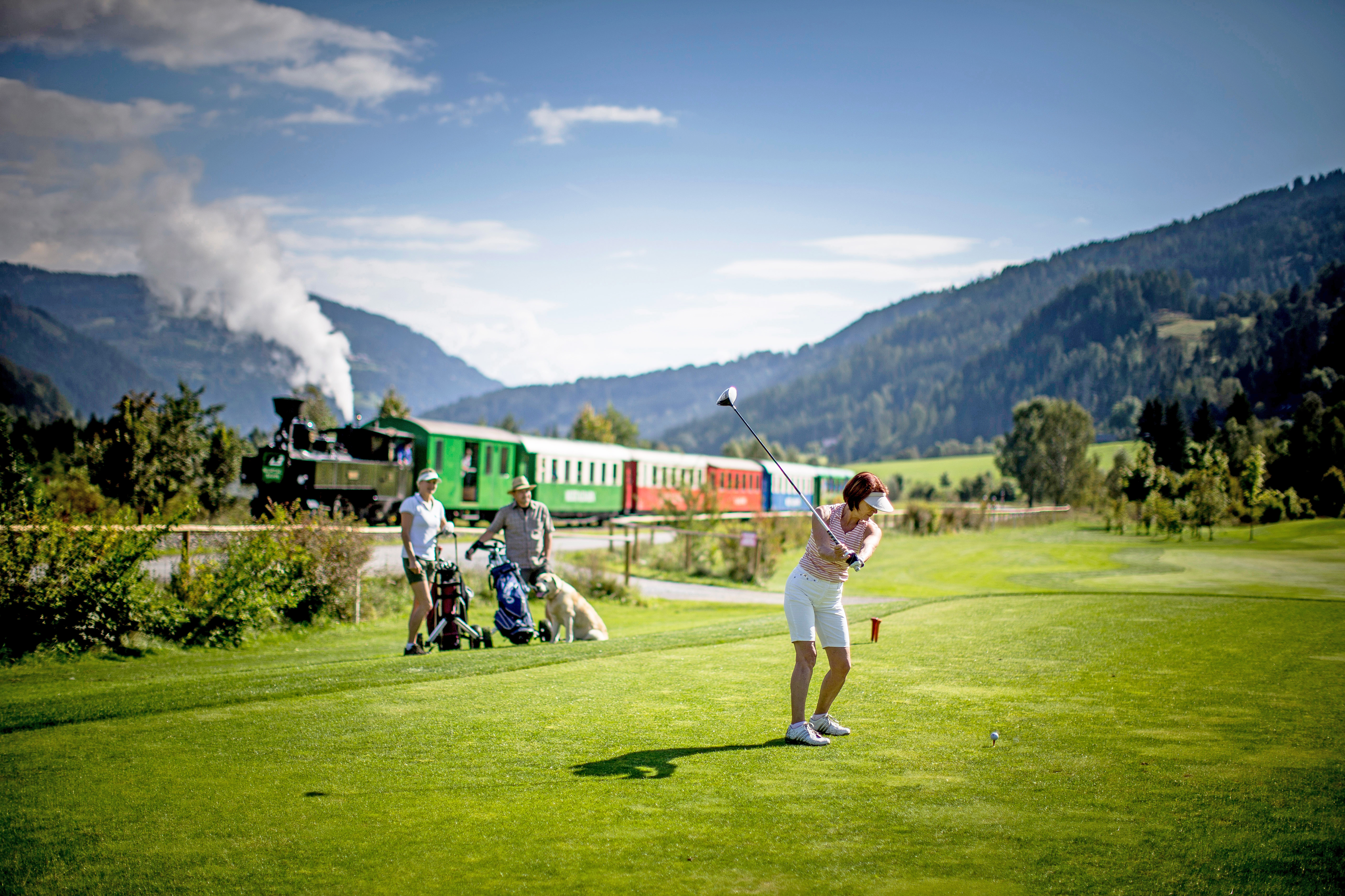 Golferin beim Abschlag am Golfplatz Murau-Kreischberg, weitere Spieler, ein Hund und die Murtalbahn vor der Bergkulisse